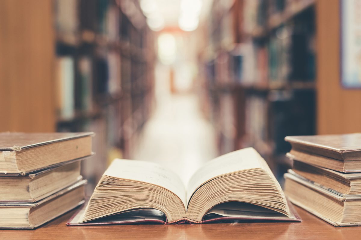 Old book in library with open textbook, stack piles of literature text archive on reading desk, and blur aisle of bookshelves in school study class room background for education learning concept