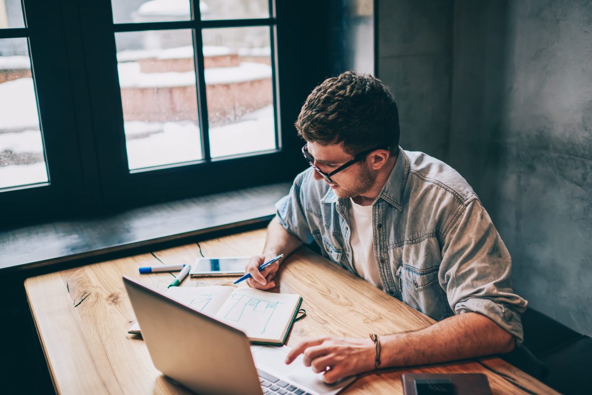 Hipster guy pondering on idea for essay sitting at wooden table with laptop device and looking on textbook, smart male freelancer spending time for distance job thinking about business indoors