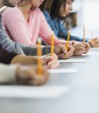 Cropped view of a multiracial group of young men and women sitting in a row at a table, writing with pencils on paper.  They are taking a test or filling out an application.  Focus is on the hand of the young man in the middle in the gray shirt.