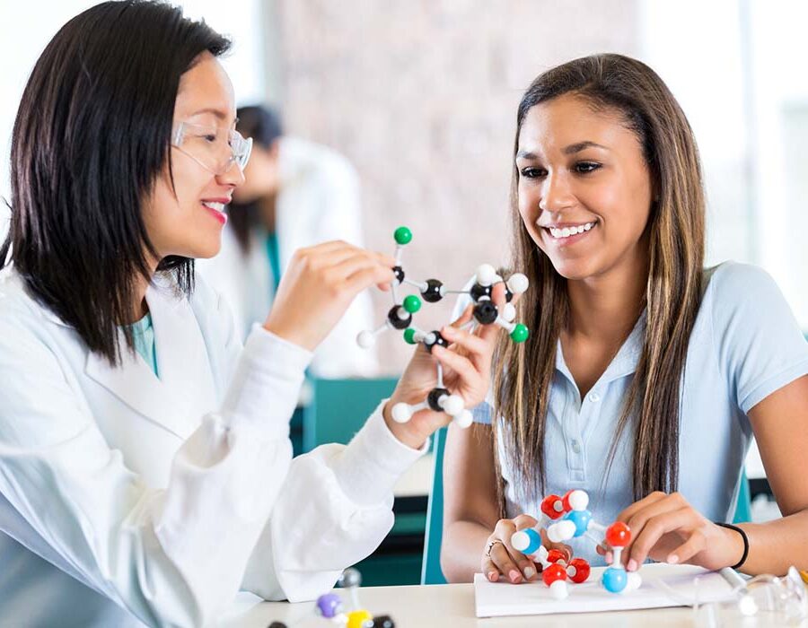 Young Asian female teacher helps teenage private high school student study molecular structure. The teacher is holding a plastic educational model. The teenage girl is interested as her teacher talks. The student is wearing a school uniform and the teacher is wearing a white lab coat.