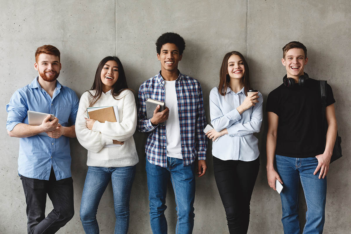 College students with books smiling to camera over grey wall, having break